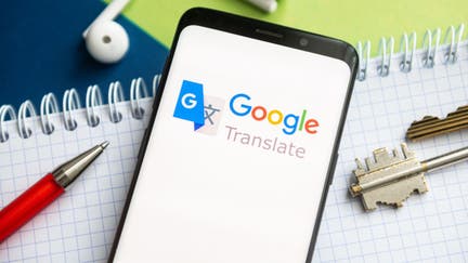 In this photo illustration, a Google Translate logo seen displayed on a smartphone with a pen, key, book and headsets in the background. 