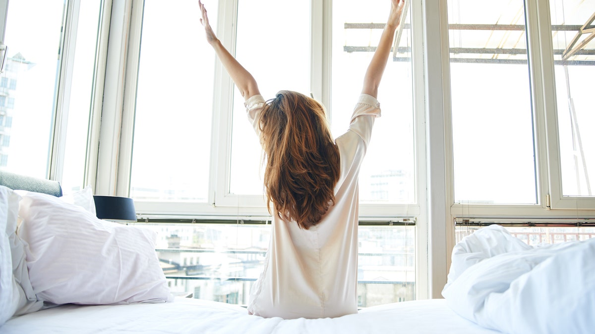 Young woman is doing morning stretching in bed, arms raised, rear view