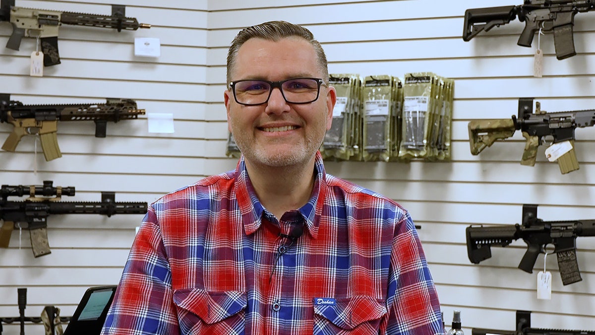 Bryan Zielinski sits in front of wall of rifles in North Idaho Arms