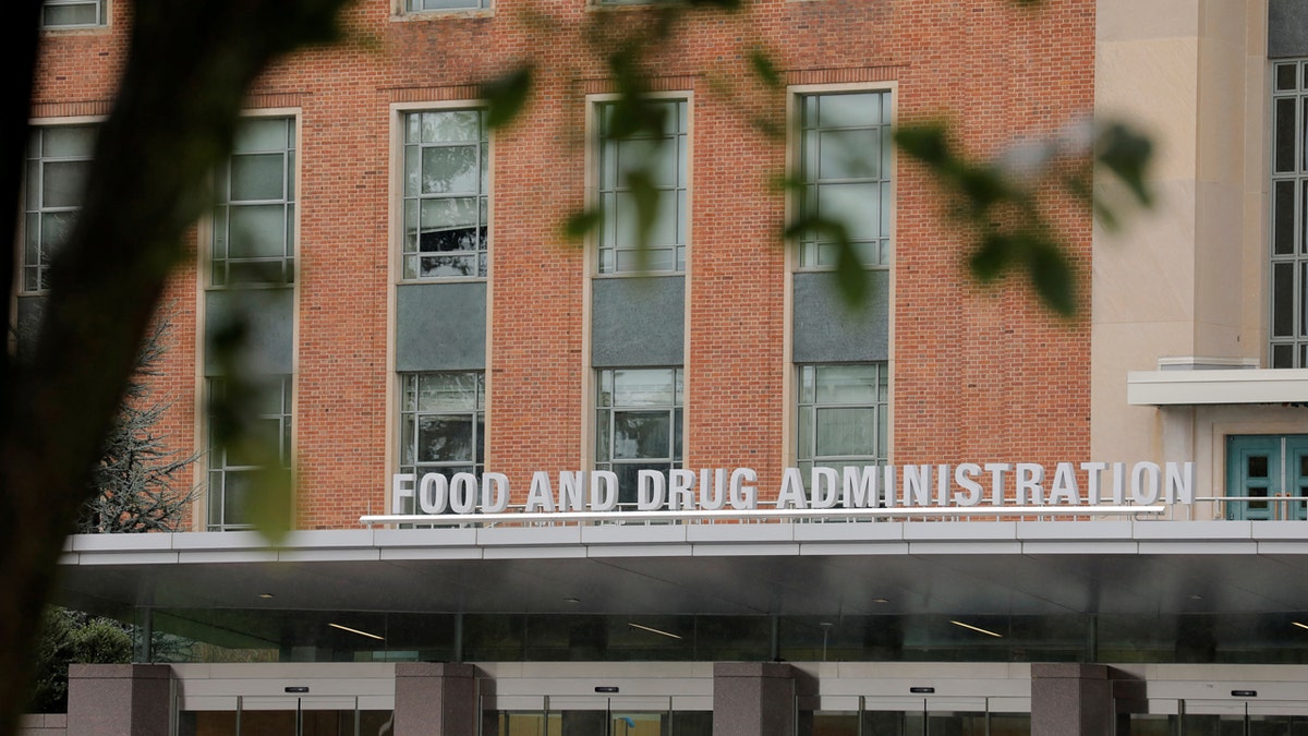 A sign in 3-D letters reading "FOOD AND DRUG ADMINISTRATION" is seen outside of the FDA headquarters with a tree out of focus in the foreground.