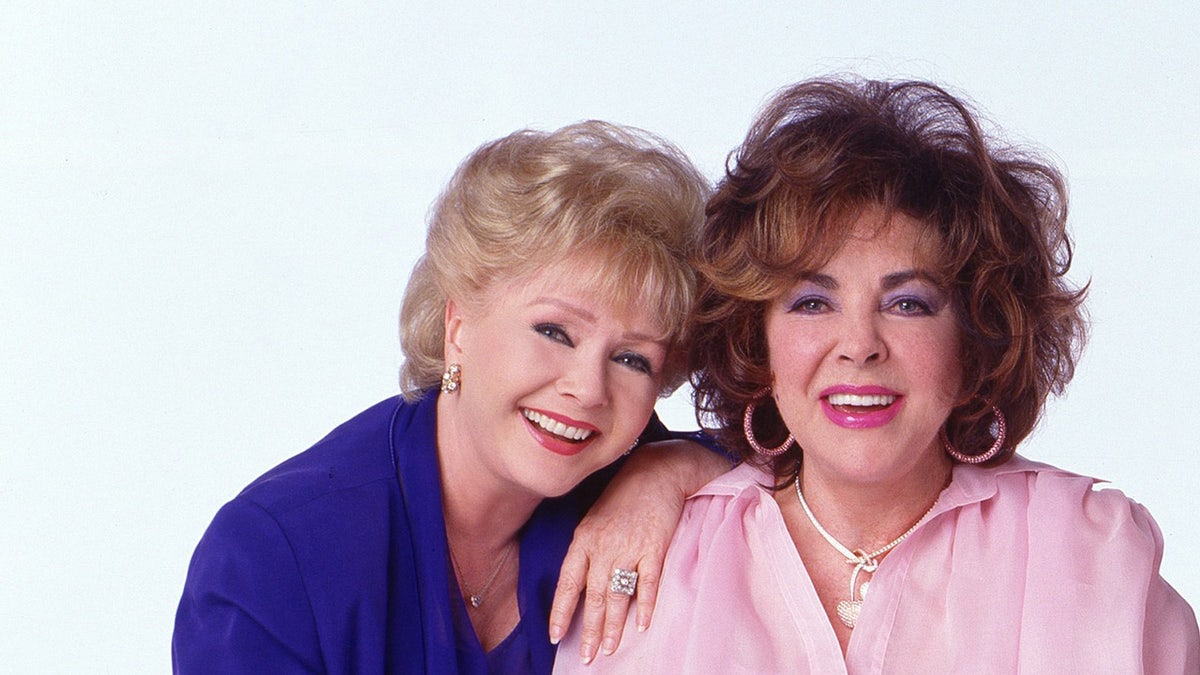 Debbie Reynolds wearing a blue blouse and smiling leaning on Elizabeth Taylor in a pink blouse and smiling.