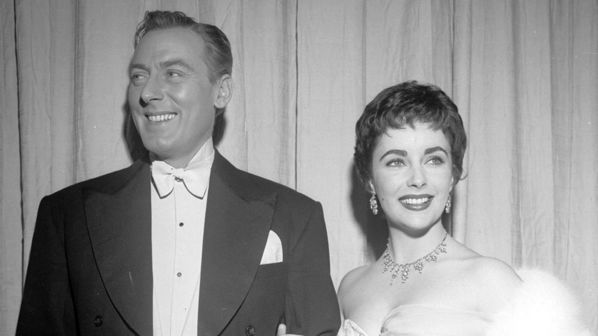 Elizabeth Taylor smiling in a glamorous gown next to Michael Wilding in a suit and white bow tie.