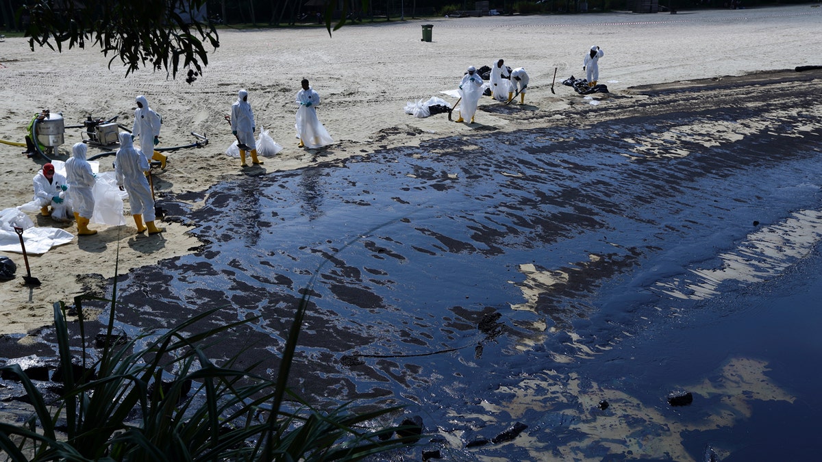 Workers wearing white suits with masks and yellow boots clean up the oil spill on the beach.