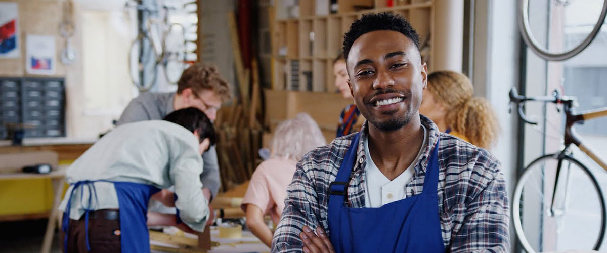 Young entrepreneur standing in his bicycle shop. Five people are working on a table behind him.