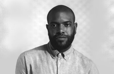 black and white photo of Black man in button up shirt in front of blurred checkered background