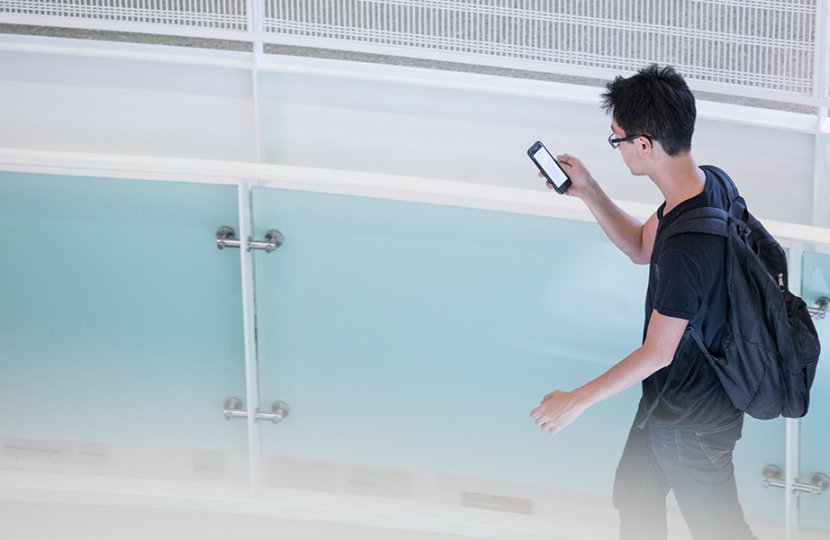 male college student wearing backpack looks at phone while walking at USC Annenberg Hall