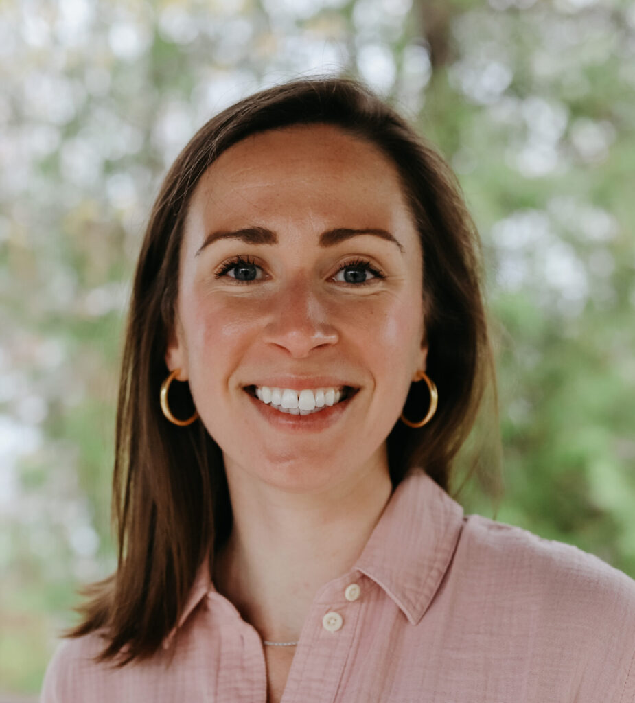 Headshot of a smiling person of light skin tone and shoulder length brown hair, wearing gold hoop earrings and a light pink button down shirt.