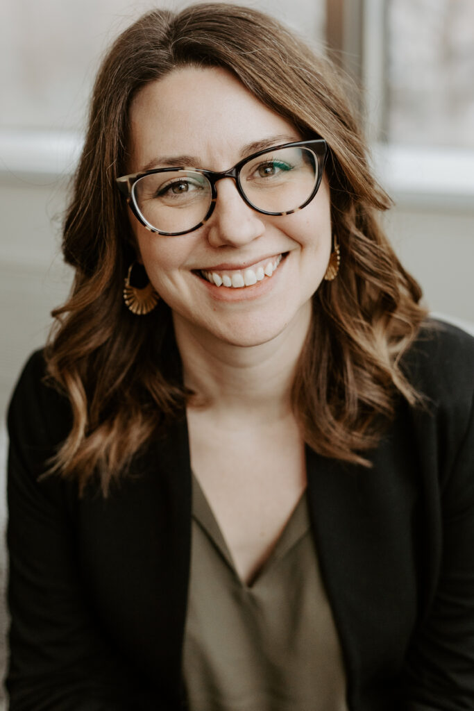 A smiling person of light skin tone and shoulder length wavy brown hair, wearing tortoiseshell rimmed glasses, earrings, and an olive green shirt under a black blazer