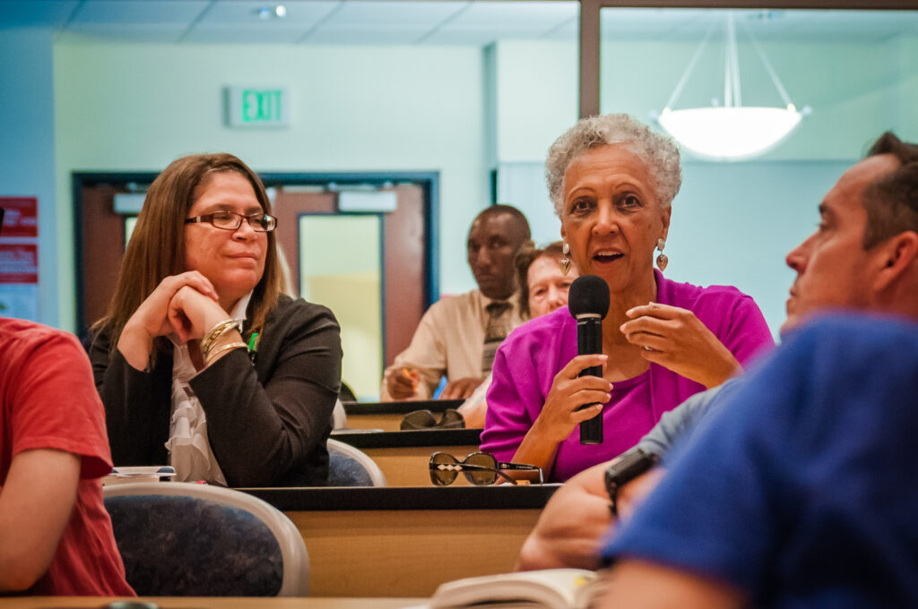 In a lecture hall, a person sitting at a table speaks into a microphone and gestures, while the people around them look and listen.