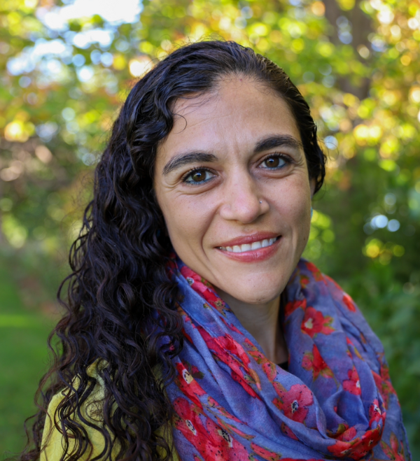 Headshot of a smiling person of medium light skin tone and long black curly hair, wearing a vibrant red and blue patterned scarf over a yellow top.