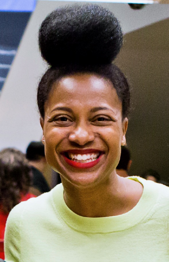 Headshot of a smiling person of medium dark skin tone, with black hair in a bun on top of their head, and wearing a bright yellow shirt.