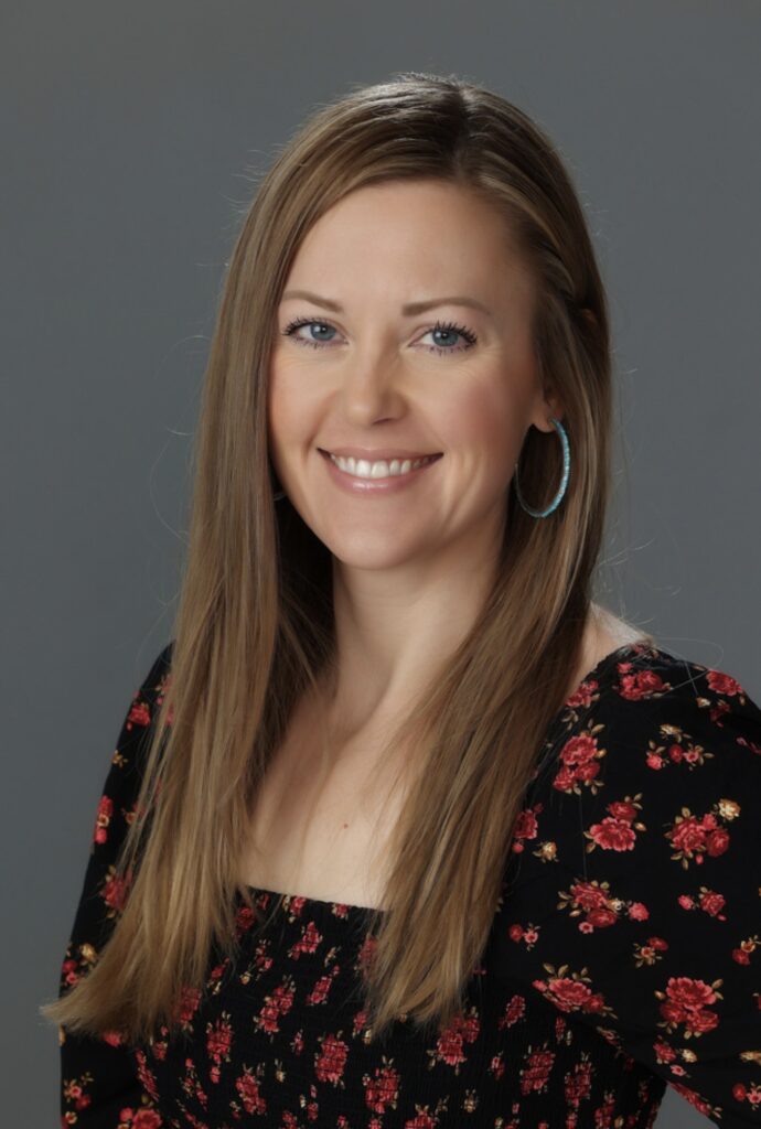 A smiling person of medium light skin tone with long light brown hair, wearing blue hoop earrings, and a black top with a red rose pattern