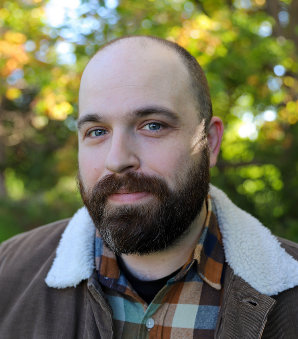 Headshot of a smiling person of light skin tone with a dark brown mustache and beard, wearing a plaid button down under a brown corduroy jacket.