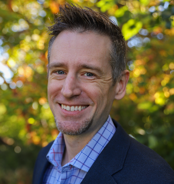 Headshot of a smiling person of light skin tone with peppered hair and a short beard, wearing a plaid button down under a navy blue blazer.