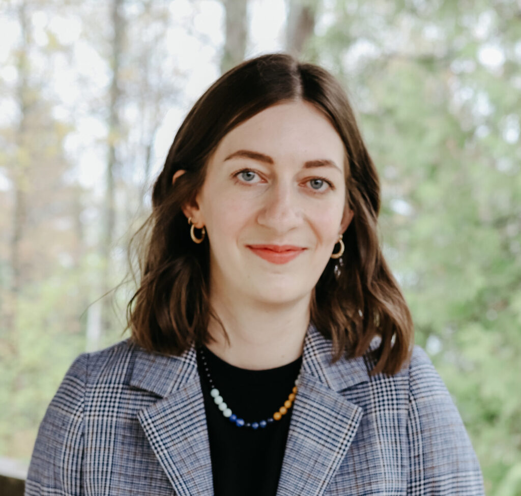 Headshot of a smiling person of light skin tone and shoulder length brown hair, wearing a vibrant beaded necklace over a black shirt and plaid blazer.