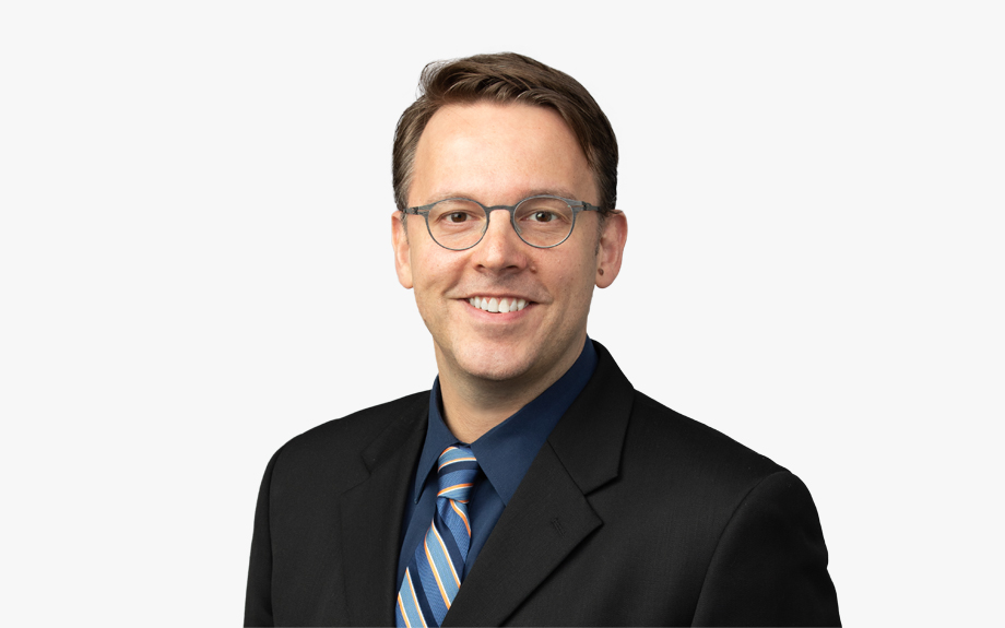 Headshot of a smiling person of light skin tone, short brown hair and dark framed glasses, wearing a striped tie, a dark blue button down shirt and a black suit jacket.