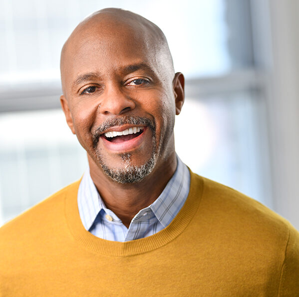 Headshot of a smiling person of medium dark skin tone, with a peppered beard and mustache, wearing a plaid shirt under a yellow sweater.