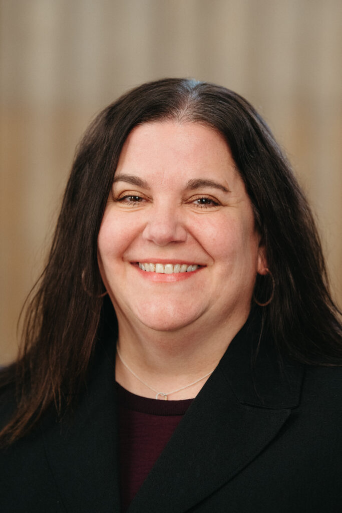 Headshot of a smiling person of light skin tone and long black hair, wearing hoop earrings, a necklace, and a maroon shirt under a black jacket.