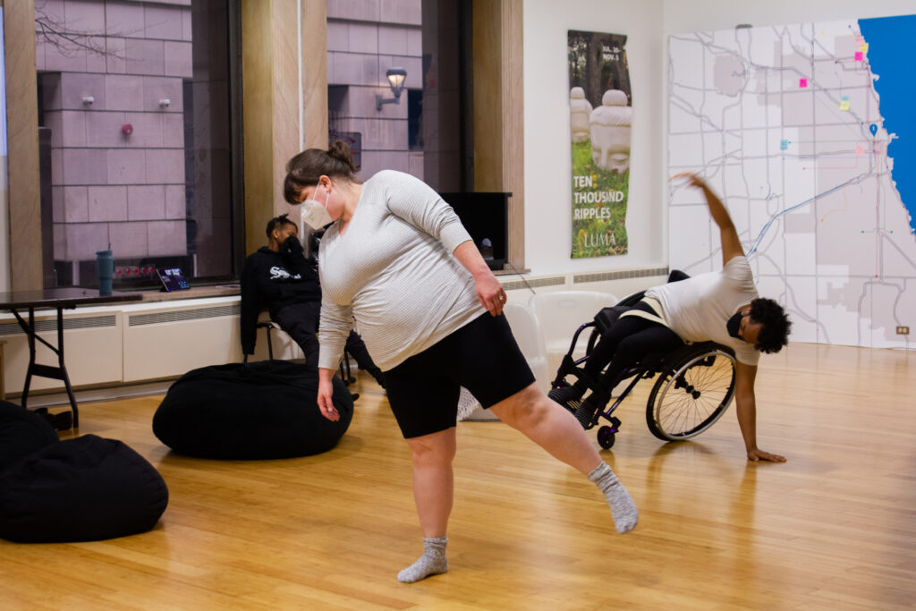 Two people dance on a hardwood floor, one is in a wheelchair and balancing on their hand touching the ground, the other is balancing on one foot and leaning to the other side