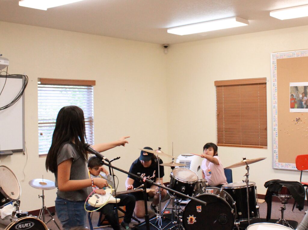 A young person on guitar and a young person on drums point to each other across the room