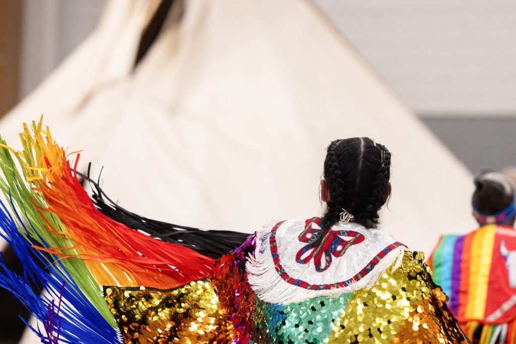A person with dark hair facing away as their colorful sequined regalia with rainbow-colored fringe flails in the air.