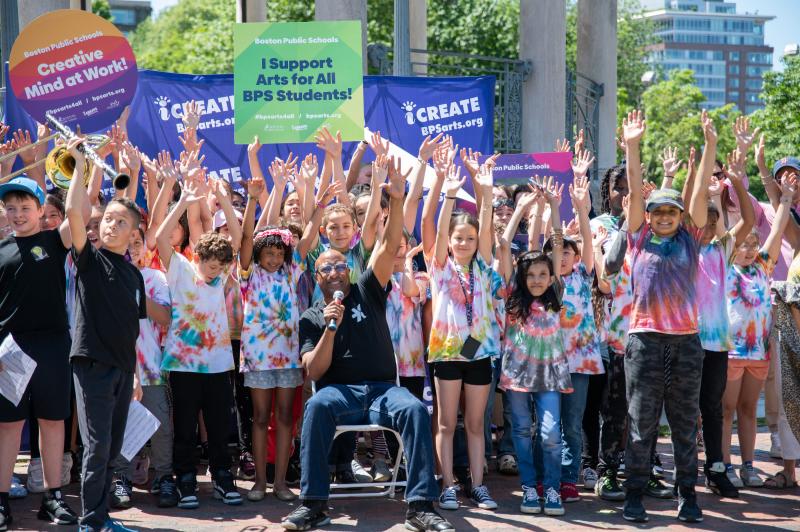 A large group of happy young people in t-shirts hold their hands up in celebration.