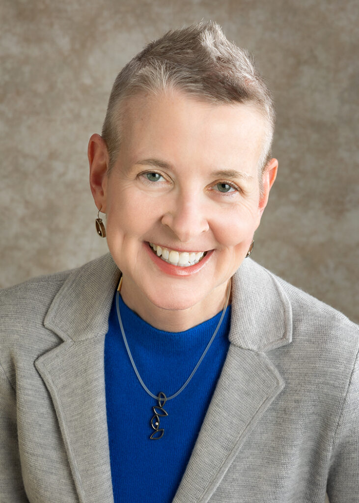 A smiling person of light skin tone with short hair wearing a blue shirt under a gray blazer.