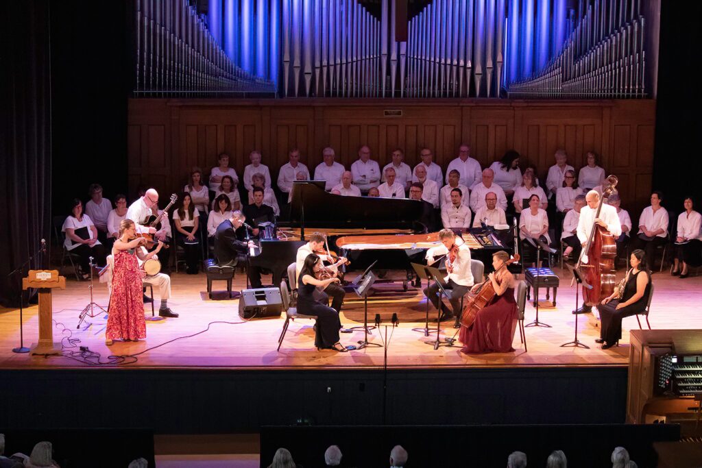 A group of musicians on stage playing the grand piano and other stringed instruments perform on a large stage. In the back of the stage are over two dozen singers seated in chairs, each wearing a white top and black bottoms.