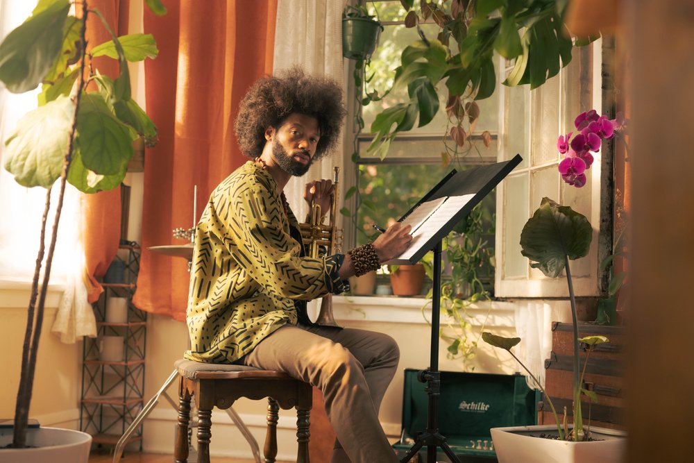 A person with medium dark skin tone and curly brown hair sitting at a music stand and holding a trumpet.