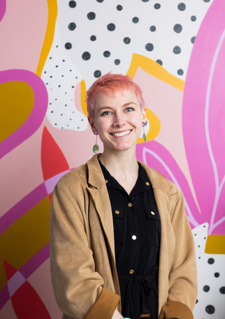 A smiling person of light skin tone with short, pink hair wearing a black blouse and tan blazer, standing in front of a pink mural.