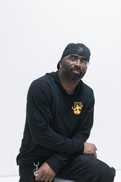 A person wearing glasses, a black hoodie, black pants, and a backwards black baseball hat sits leaning with their arms crossed in front of them in front of a white background.