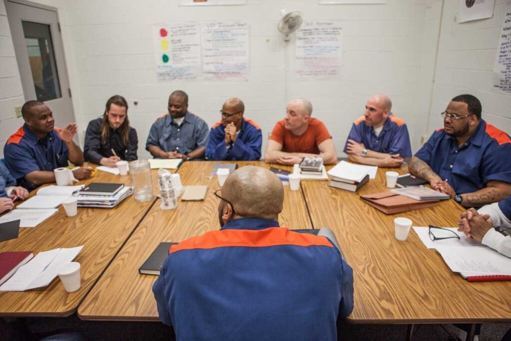 A group of people, some of whom wear orange and blue jumpsuits, sit in a half circle around a large wooden table. On the white brick wall behind them are large pieces of paper written on with colorful market. In front of them on the table are cups, water bottles, notebooks, and glasses. One person is writing, the rest are turned towards someone on the right side of the table who is speaking.