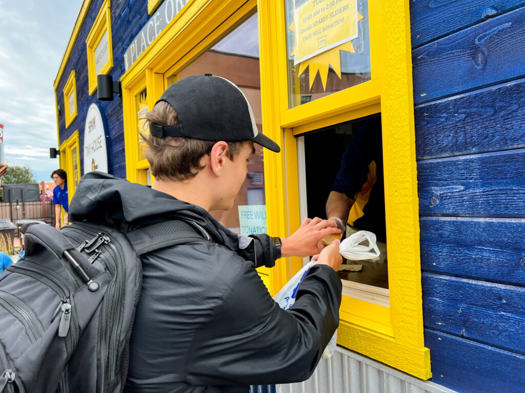 A person in a black raincoat receiving a slider from a person inside a tiny house through a yellow window.