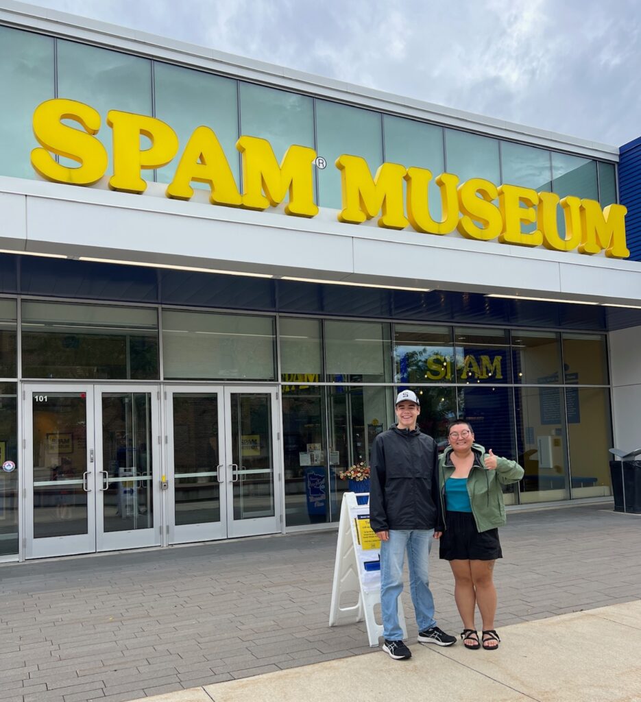 Two smiling people standing in front of a building with big yellow signage reading, "SPAM Museum"