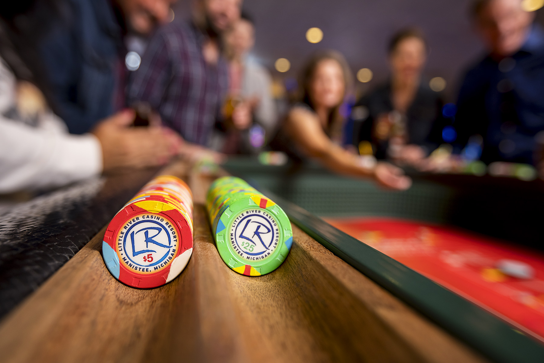 Closeup of Little River Casino Resort’s red and green casino chips stacked sideways. In the background, gamers roll dice on a red-felt craps table.  