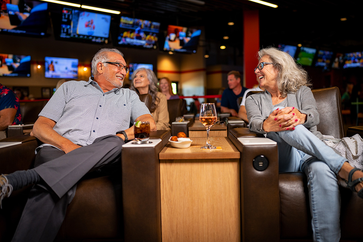 A smiling elderly couple sitting and enjoying some wine and a cocktail inside the sports bar at Little River Casino Resort. 