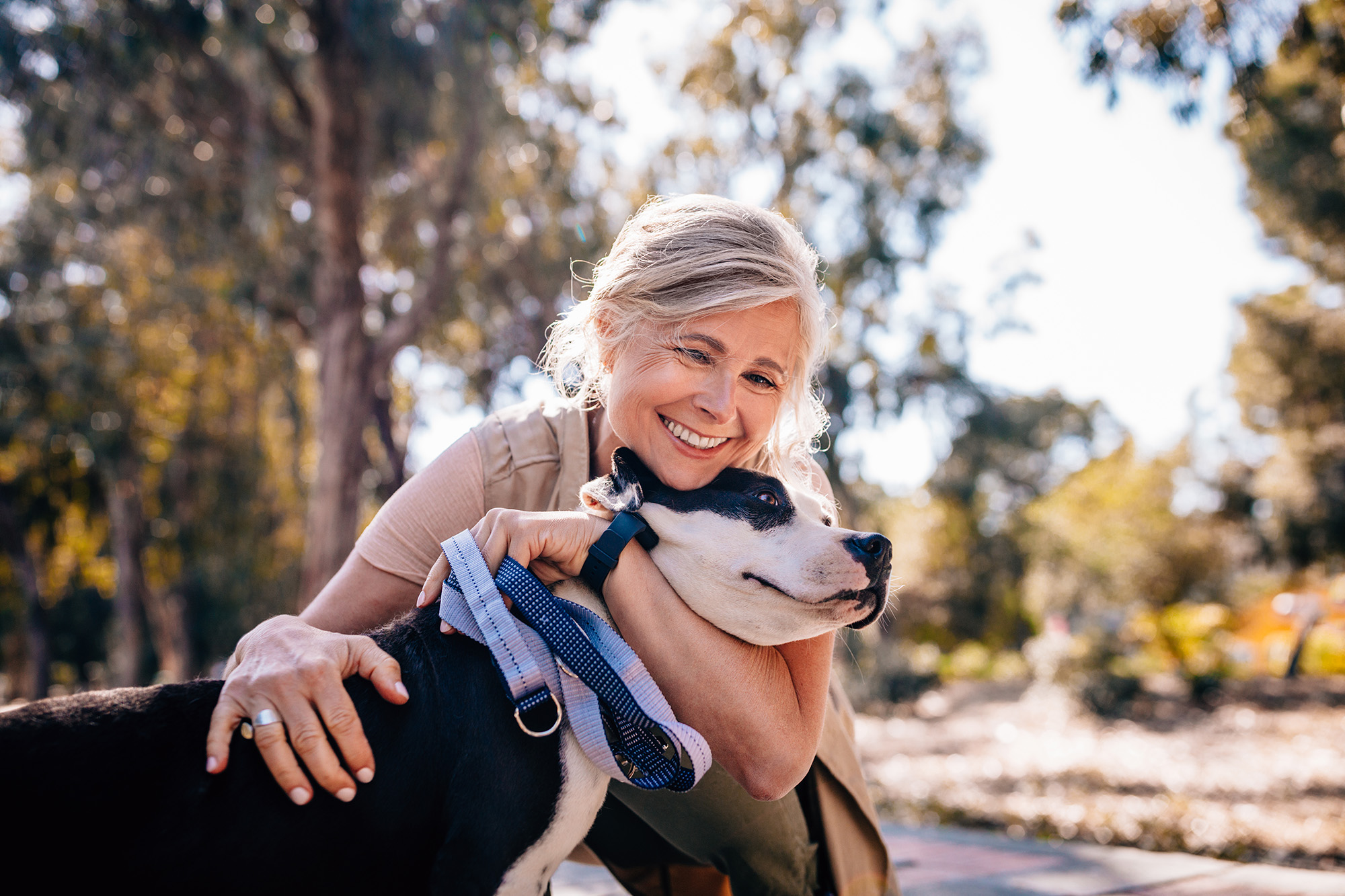 Happy senior woman enjoying walk in nature and embracing pet dog in forest park.