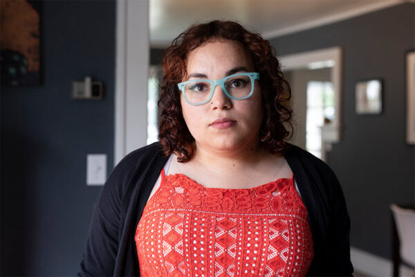 Woman with short red hair, wearing a red dress and blue glasses, looks at the camera in a grey room.