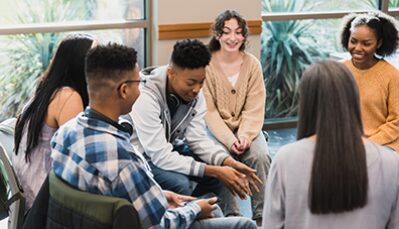 A multiracial group of young people chat, lending each other support. The smile as a young Black man leads the discussion.