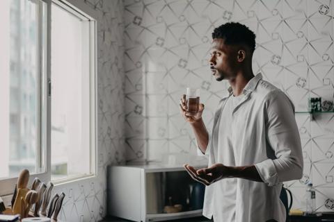 Person standing in kitchen holding glass of water in one hand and medication in the other