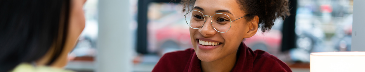 Two women sit across a table from each other while smiling. One woman is wearing a yellow shirt and the other is wearing glasses and a red shirt.