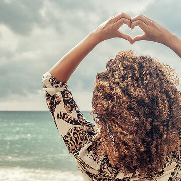 A woman with beautiful curly hair at the beach, forming a heart with her fingers above her head