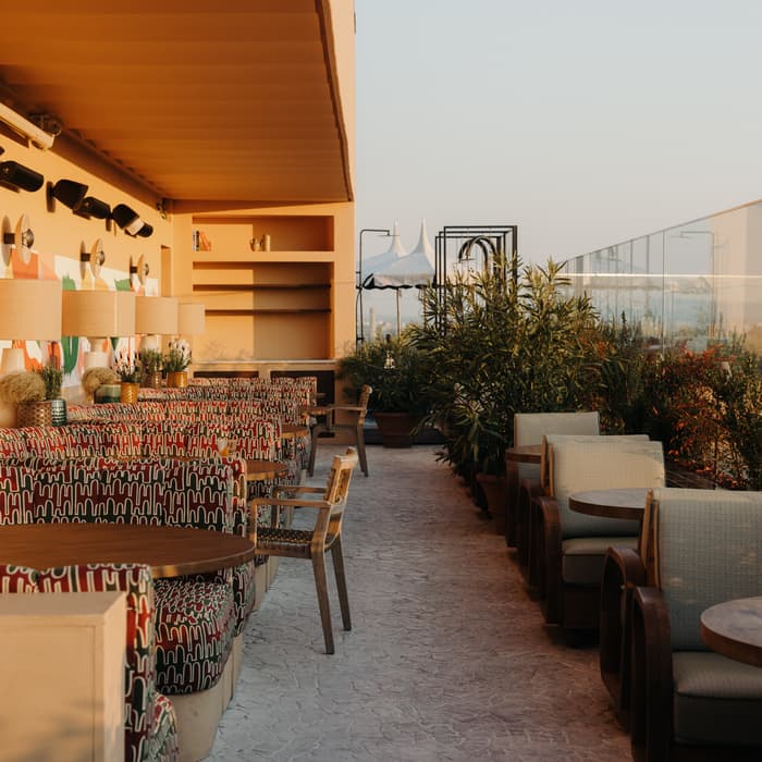 Rooftop restaurant features patterned chairs and round wooden tables set along a decorative wall with lamps and plants, offering an airy and lush ambiance with city views.