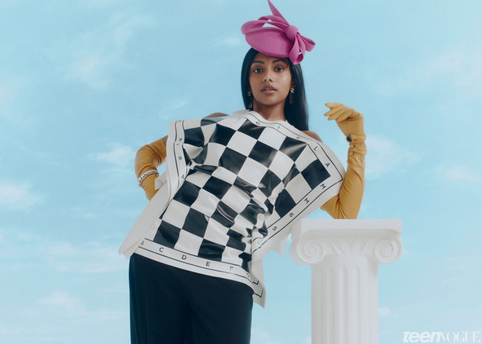 Charithra Chandran wearing a top that looks like a chess board yellow gloves and a pink headpiece. She's leaning on a...