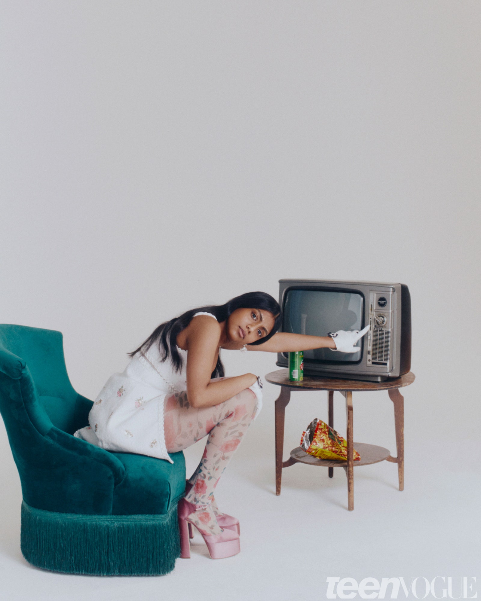 Charithra Chandran wearing a white dress floral tights and platform heels sitting on a green chair and reaching toward...