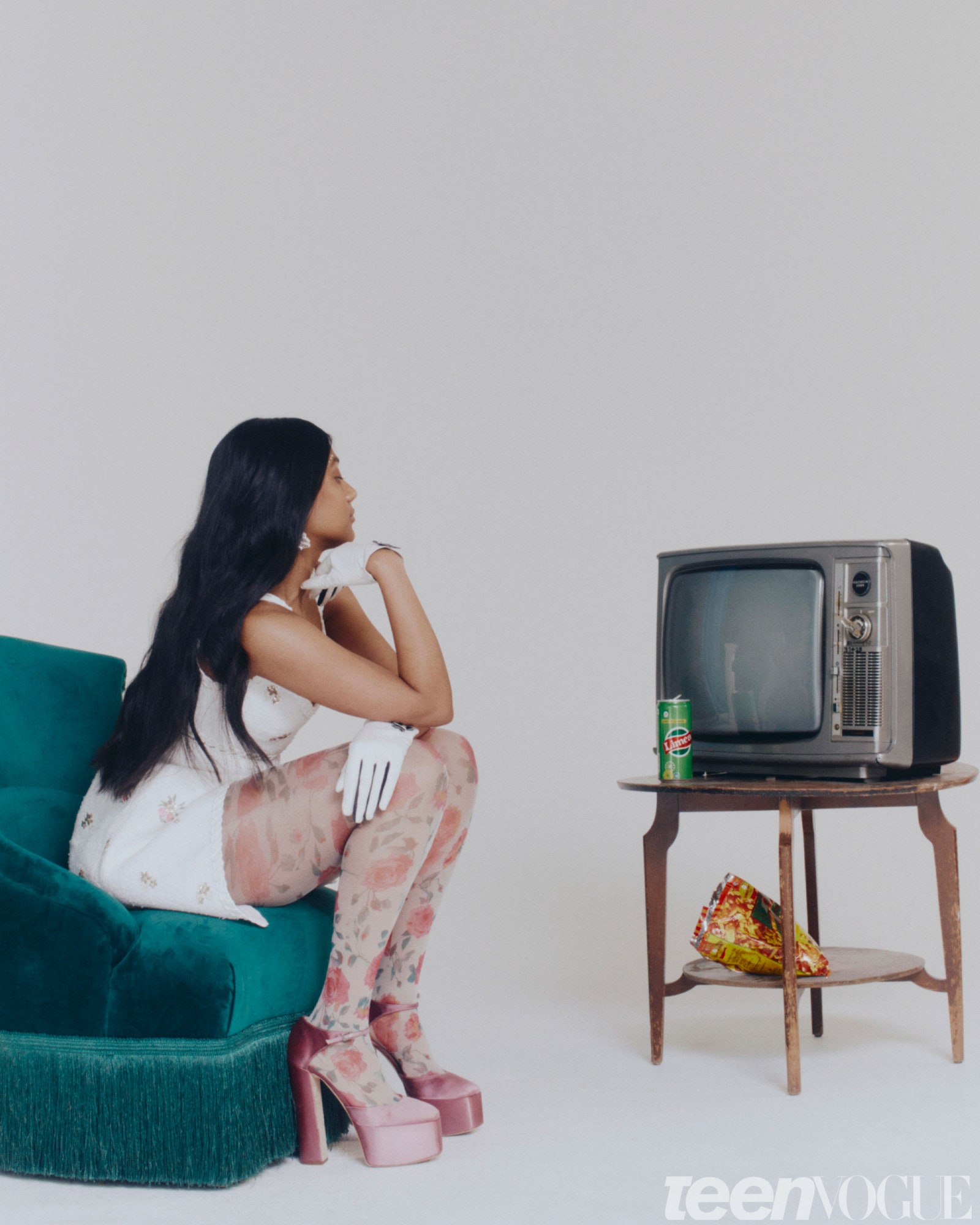 Charithra Chandran wearing a white dress floral tights and platform heels sitting on a green chair in front of an old...