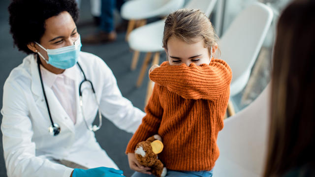 Little girl coughing into elbow while being at medical clinic. 