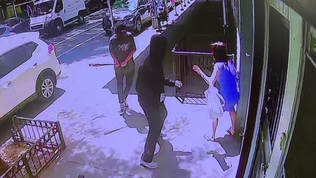 A masked man with a baseball bat and a second masked man approach a woman on a New York City street. 