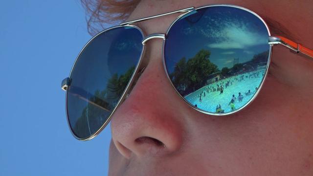 A swimming pool full of people reflected in a lifeguard's sunglasses. 