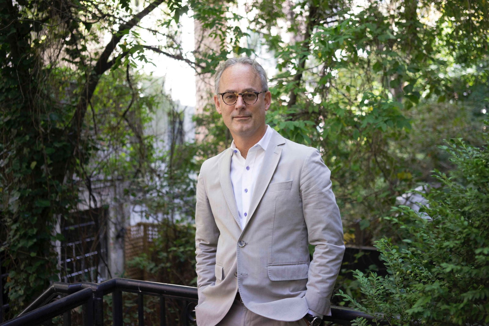 A photo of Amor Towls in a grey suit and glasses in front of elafy trees